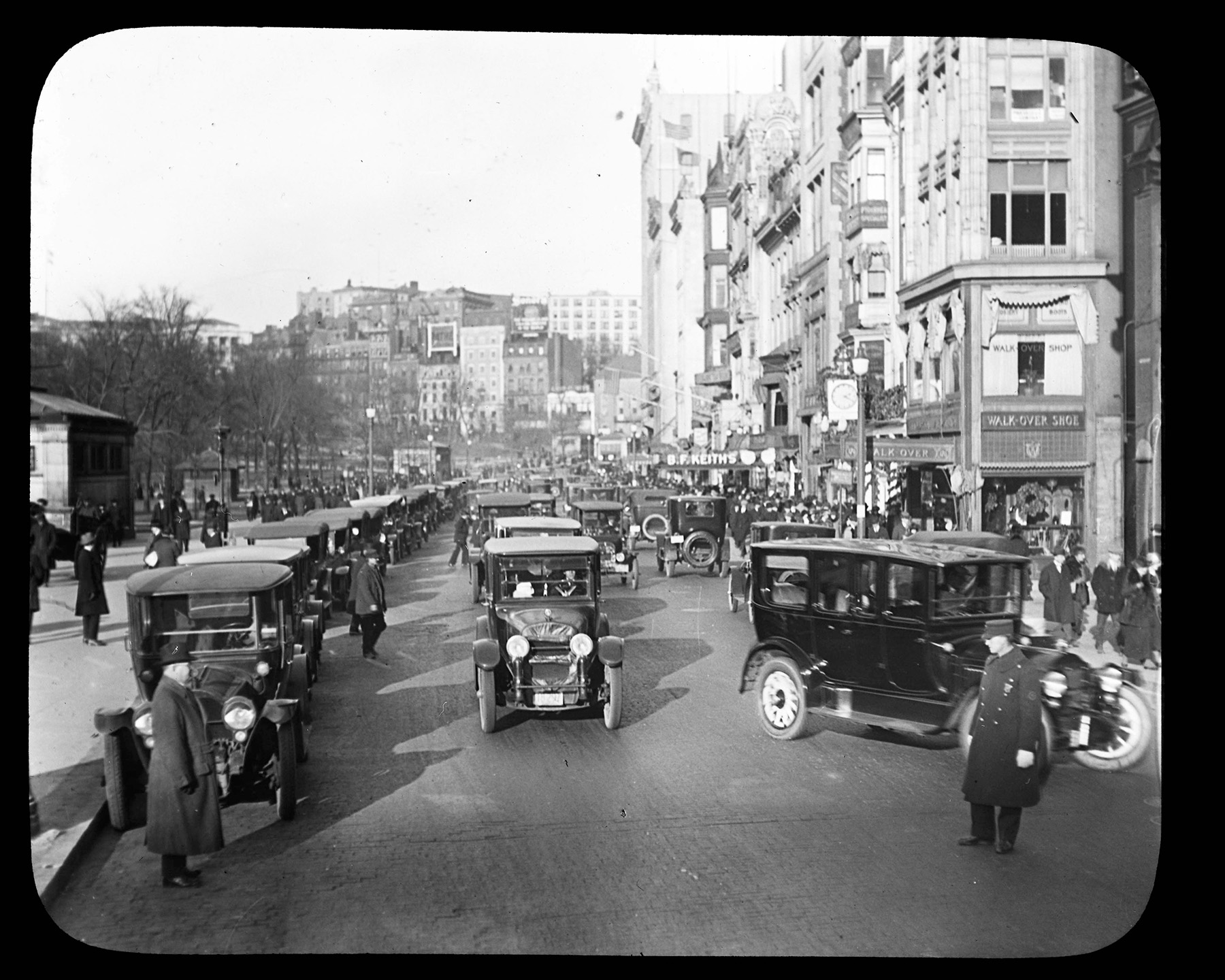 Black and white photograph depicting a street crowded with vehicles. Two men can be seen in the foreground directing traffic.