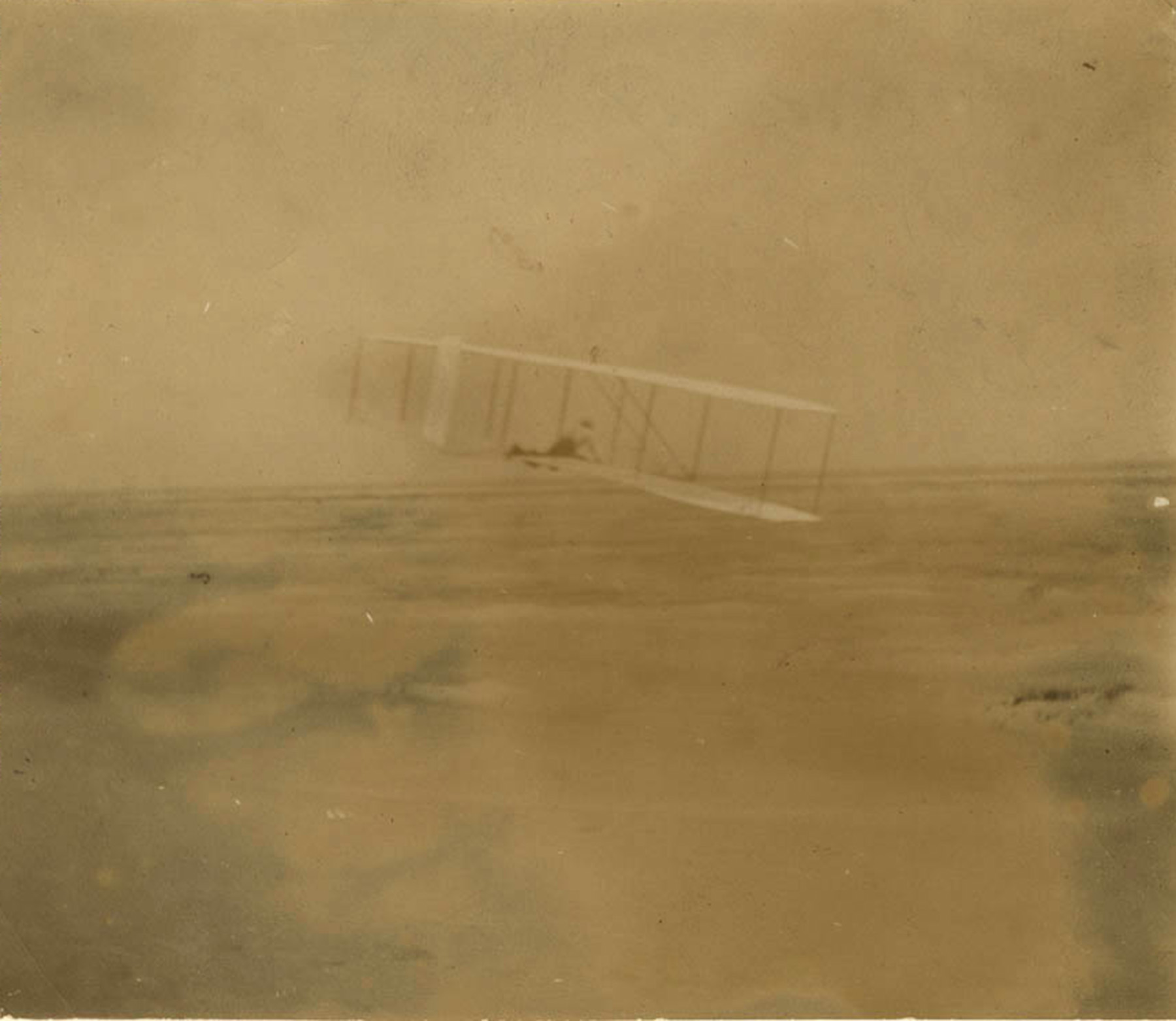 Yellowed photograph showing a glider with two wings and a person lying on the middle of it while in mid-flight