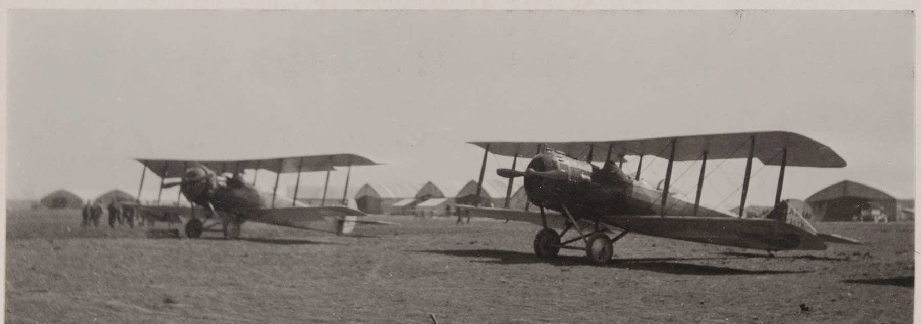 Black and white photograph of two small plane. Several tents and a group of soldiers can be seen in the background.