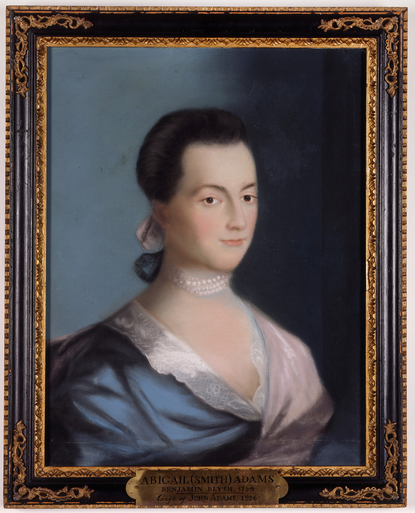 A framed portrait of a young woman with dark hair. She wears a blue dress with lace around the neckline, a pink wrap, a pearl choker necklace, and a pink ribbon in her hair.