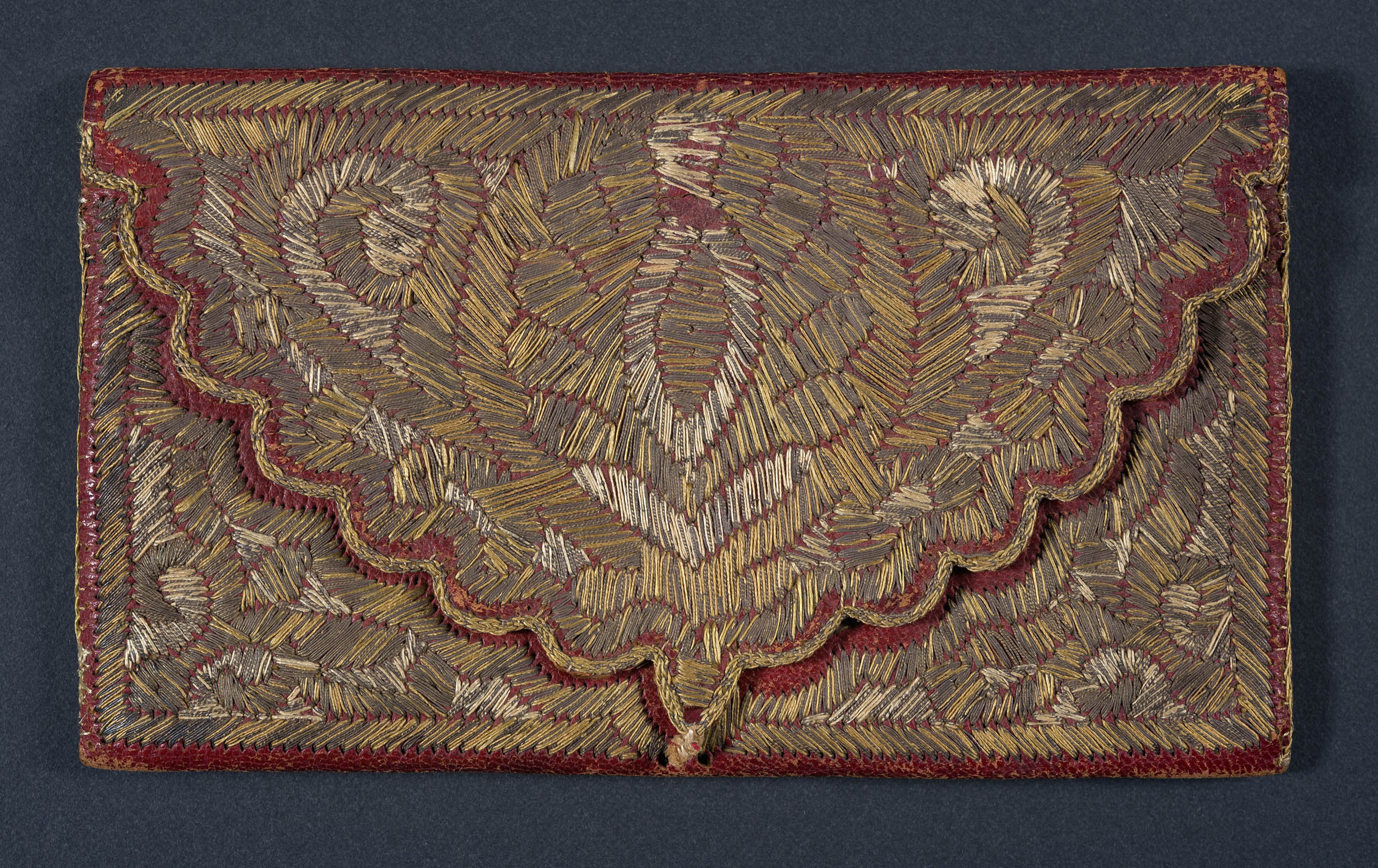 rectangular red leather wallet embroidered all over with linen and metallic threads; the folded over flap has scalloped edges.