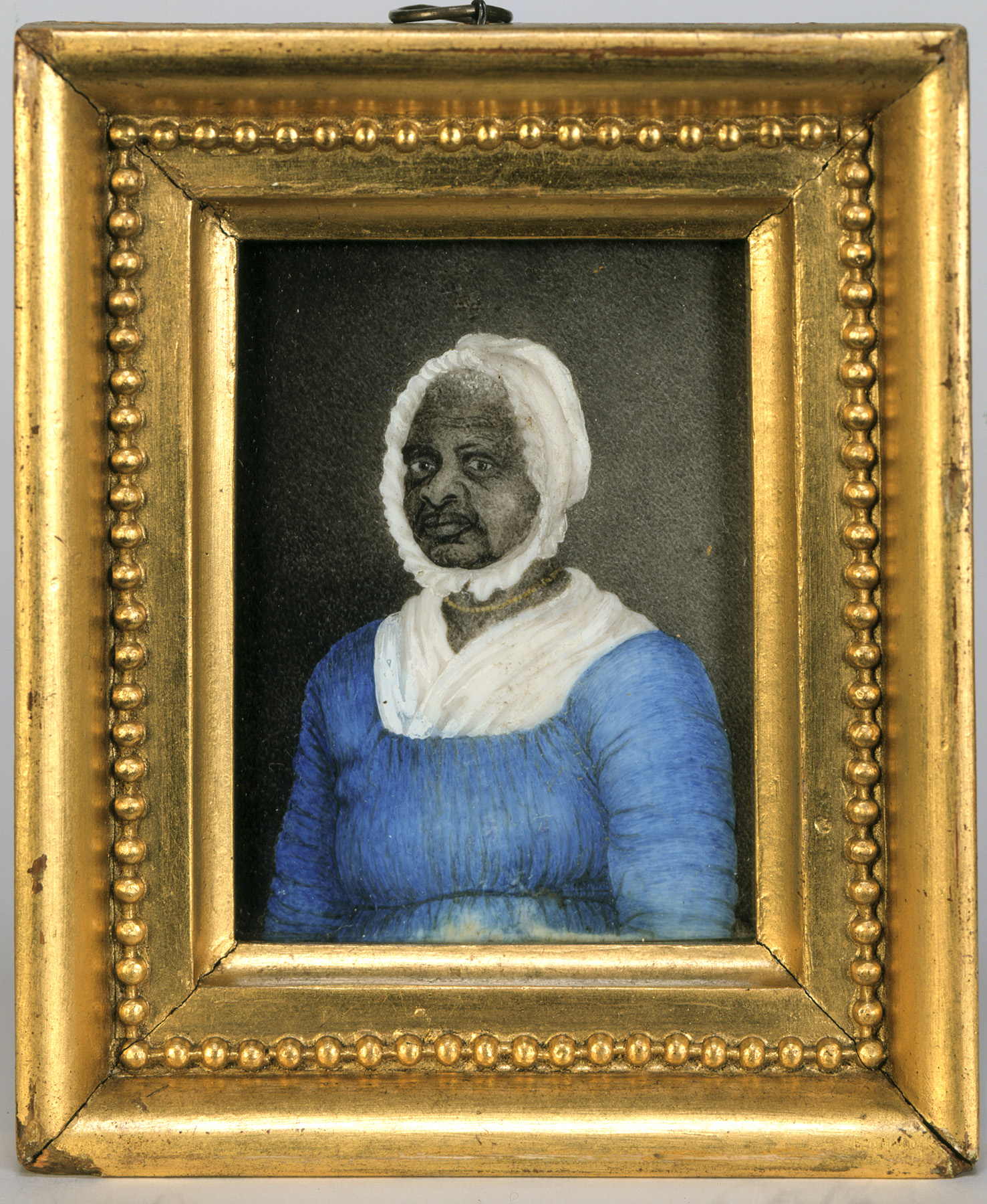 A framed portrait of a Black woman wearing a blue dress with drawstrings at the neck and waist, a white fichu tucked into her dress at the neck, a white cap, and a gold beaded necklace.