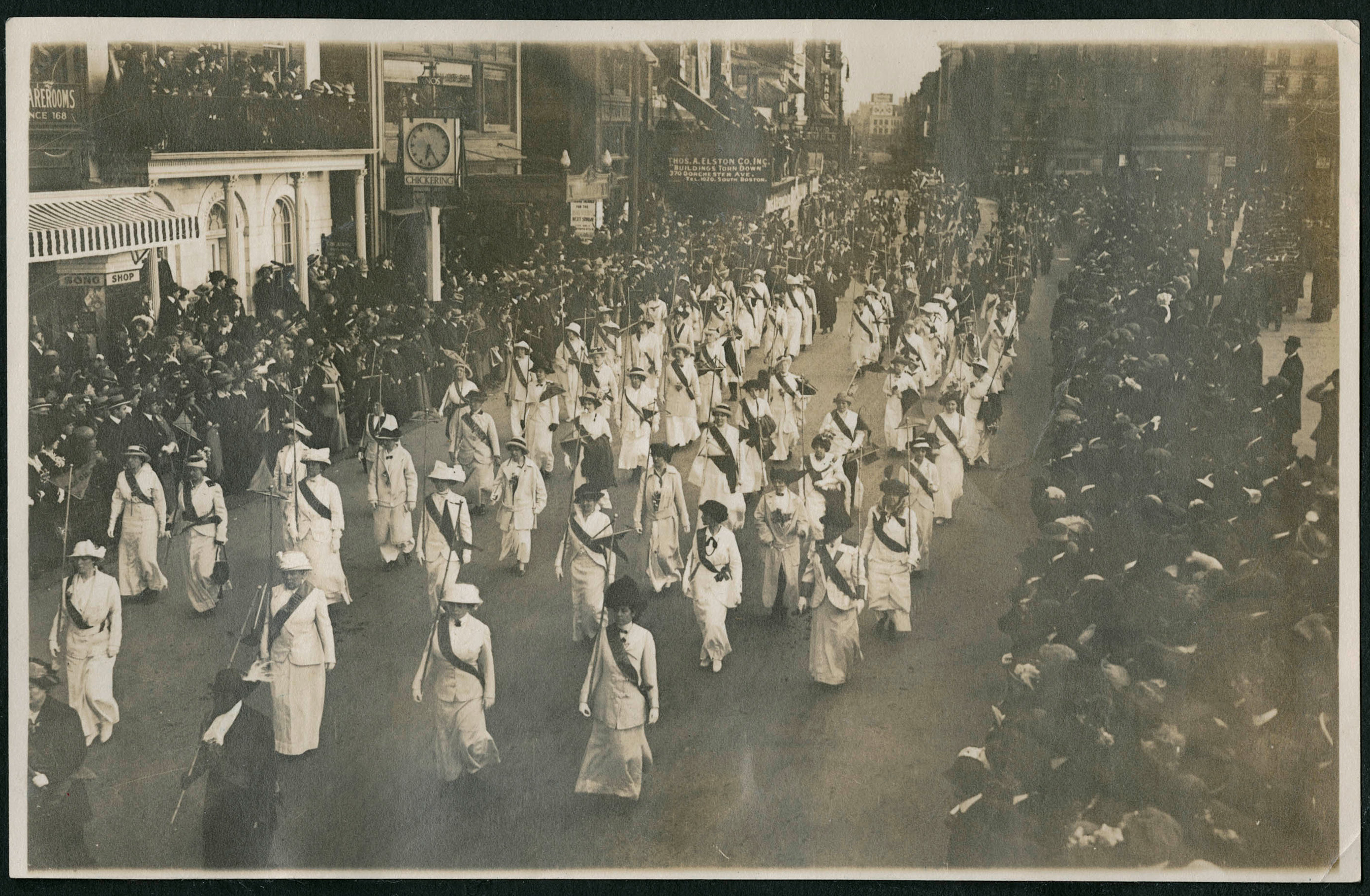 A black and white photo of a number of women marching in the street, wearing matching white outfits and dark sashes while crowds look on